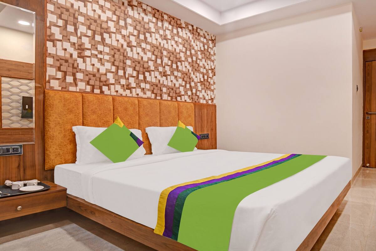 A modern hotel room with a wooden headboard, a large bed with a green and purple blanket, and a wooden wall panel.