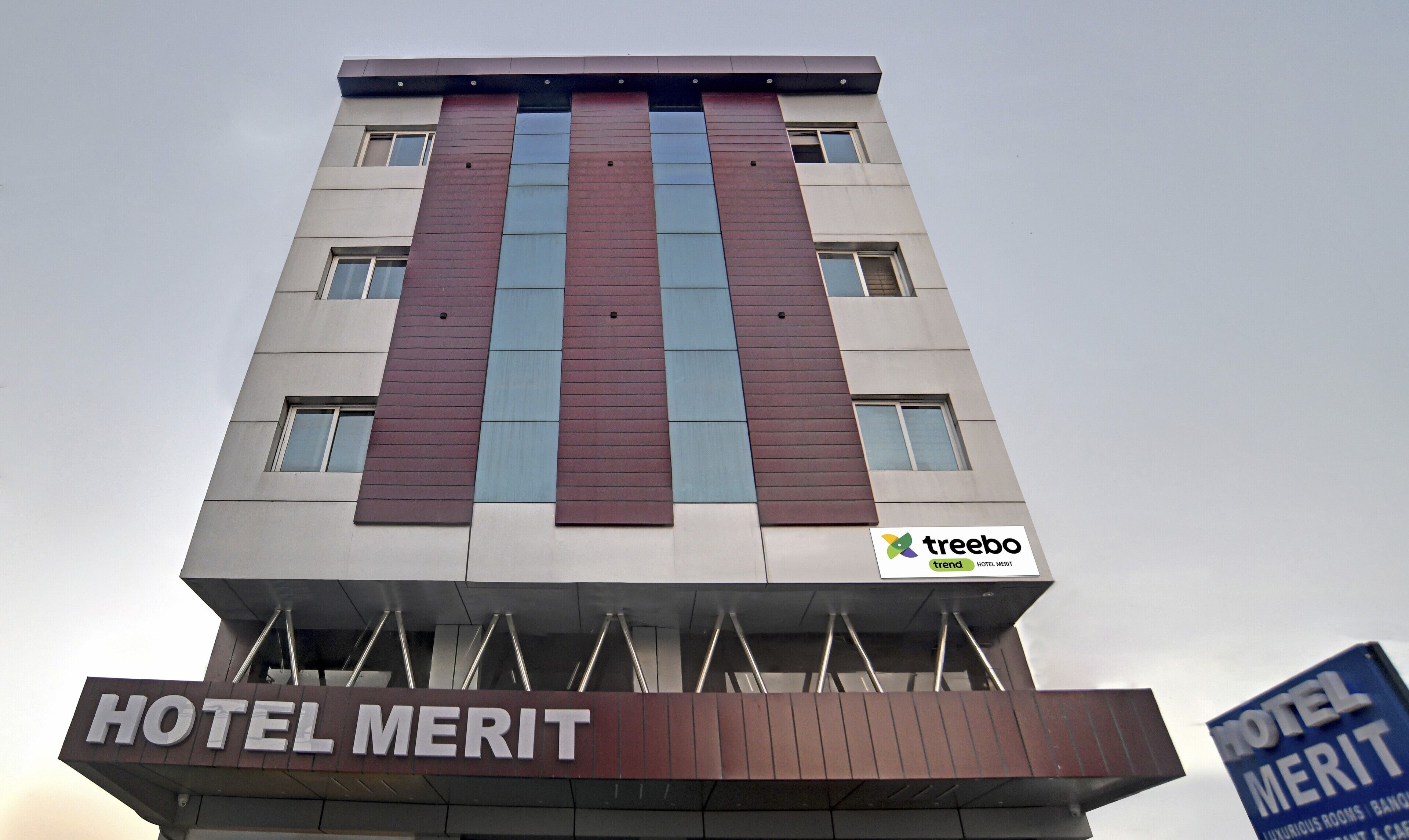 Un hôtel appelé HOTEL MERIT, doté d’un design moderne, avec une enseigne bien visible et un logo indiquant qu’il fait partie du réseau TREEBO TREND HOTEL.