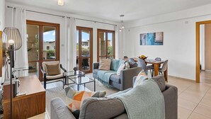 A modern living room with a sofa, armchairs, a coffee table, and a dining area with a table and chairs. There are large windows and a view of the outside. - Apartment BH01 - Anthos (Kouklia)