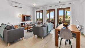 A modern living room with a sofa, a coffee table, and a TV. There is a dining area with a table and chairs. The room has large windows and a view of the outside. - Apartment BH01 - Anthos (Kouklia)