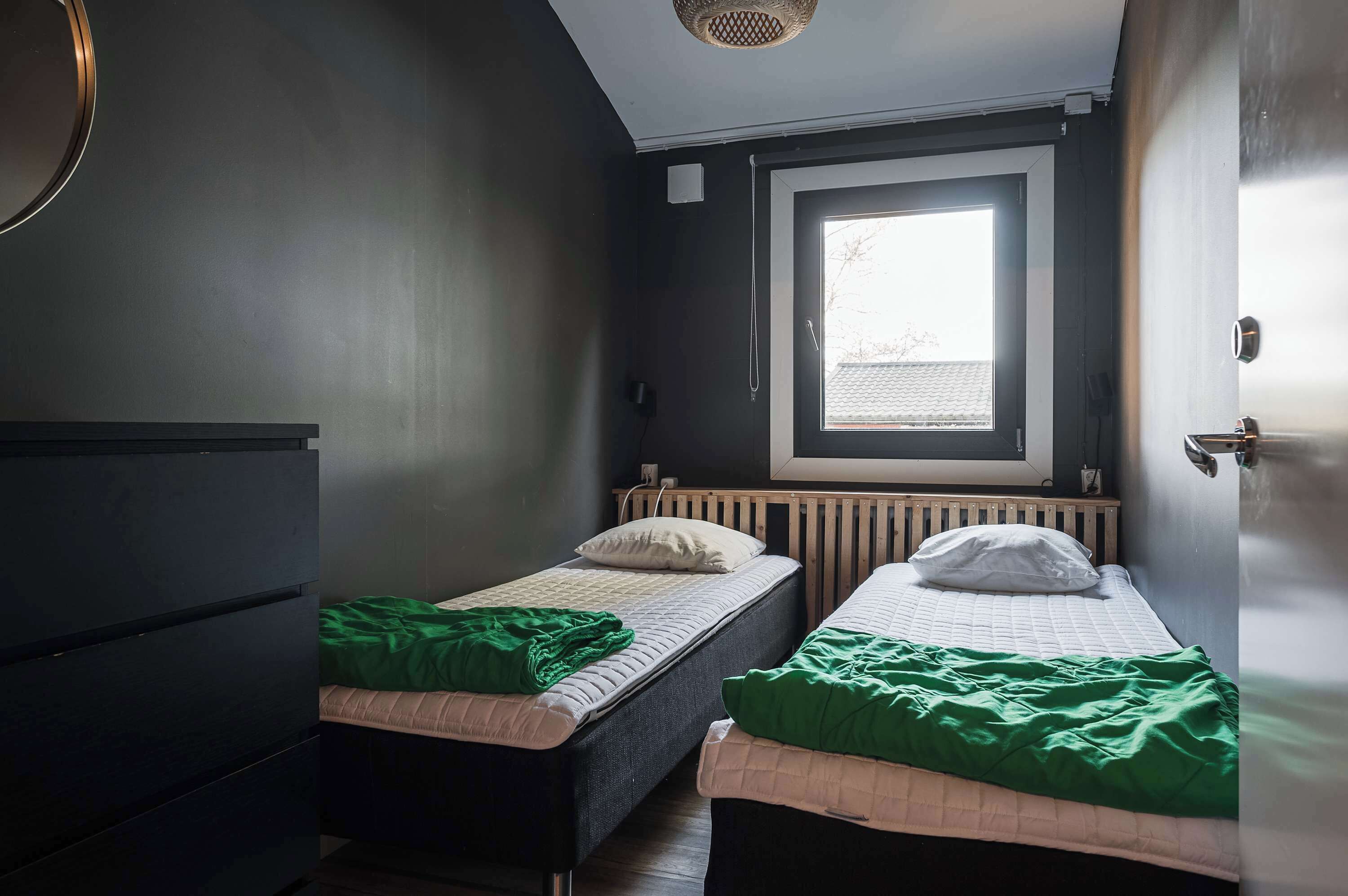 Une petite pièce peu éclairée, avec deux lits simples, une commode sombre et une fenêtre avec des stores.