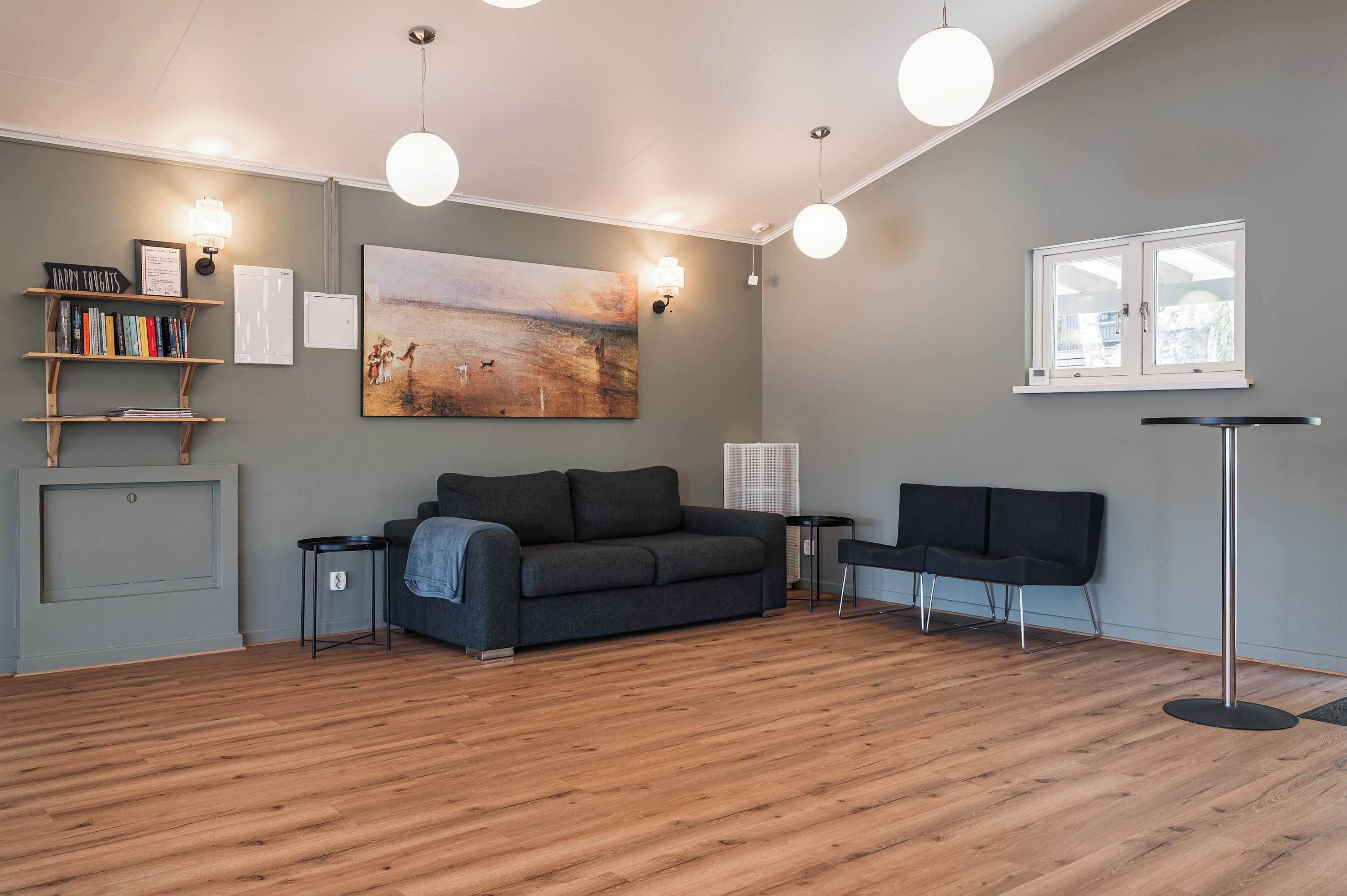 Un salon moderne avec un canapé gris, un fauteuil noir, un plancher en bois, un tableau et une bibliothèque.