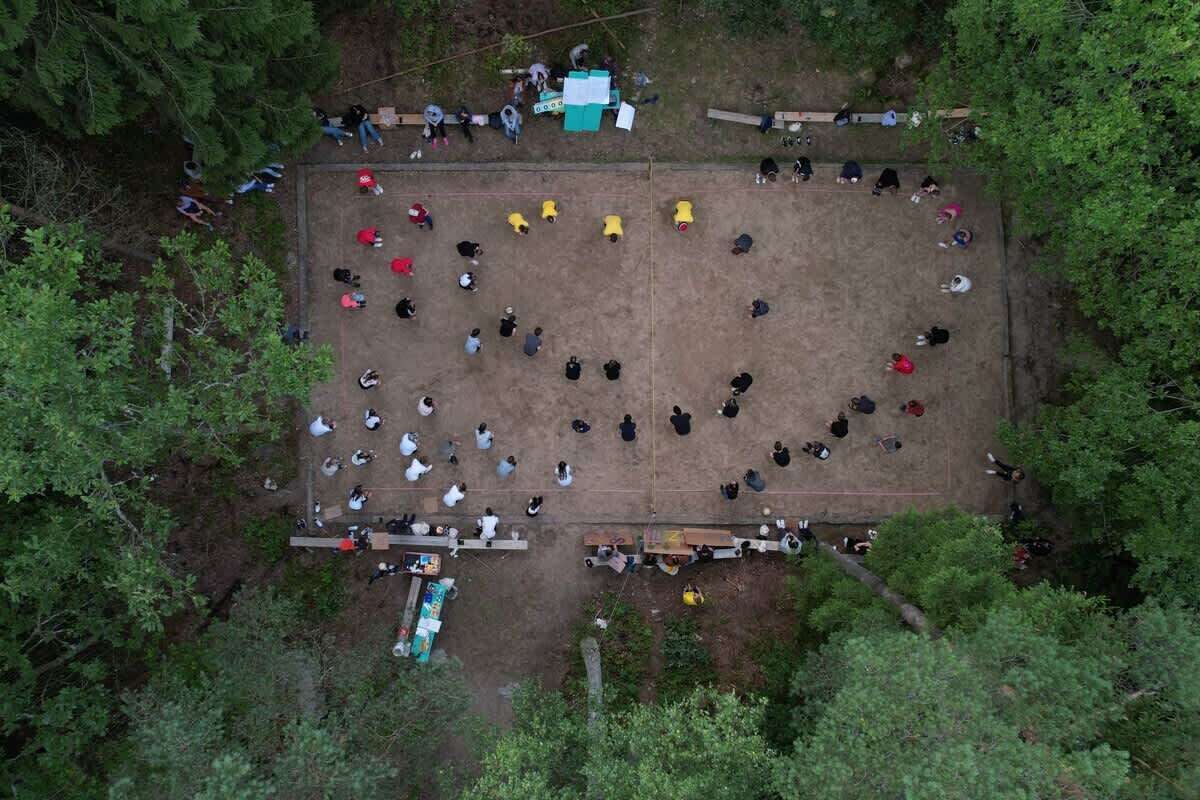 Vista aérea de un espacio exterior rectangular rodeado de árboles, con personas reunidas en un círculo central, bancos y una pequeña construcción con techo verde.