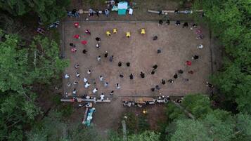 Vista aérea de un espacio exterior rectangular rodeado de árboles, con personas reunidas en un círculo central, bancos y una pequeña construcción con techo verde.