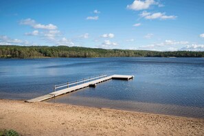 A sandy beach with a wooden dock extending into a calm lake, surrounded by a forested area.