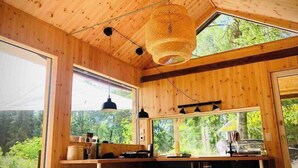 A wooden cabin interior with large windows, a hanging light fixture, and a kitchen area. - The Villa – Lake Escape w/ Sauna, Jacuzzi & Boats (Vegby)