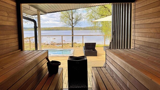 A wooden deck with a hot tub, a black grill, and a view of a lake. - The Villa – Lake Escape w/ Sauna, Jacuzzi & Boats (Vegby)