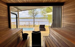 Eine Holzterrasse mit Whirlpool, einem schwarzen Grill und Blick auf einen See.