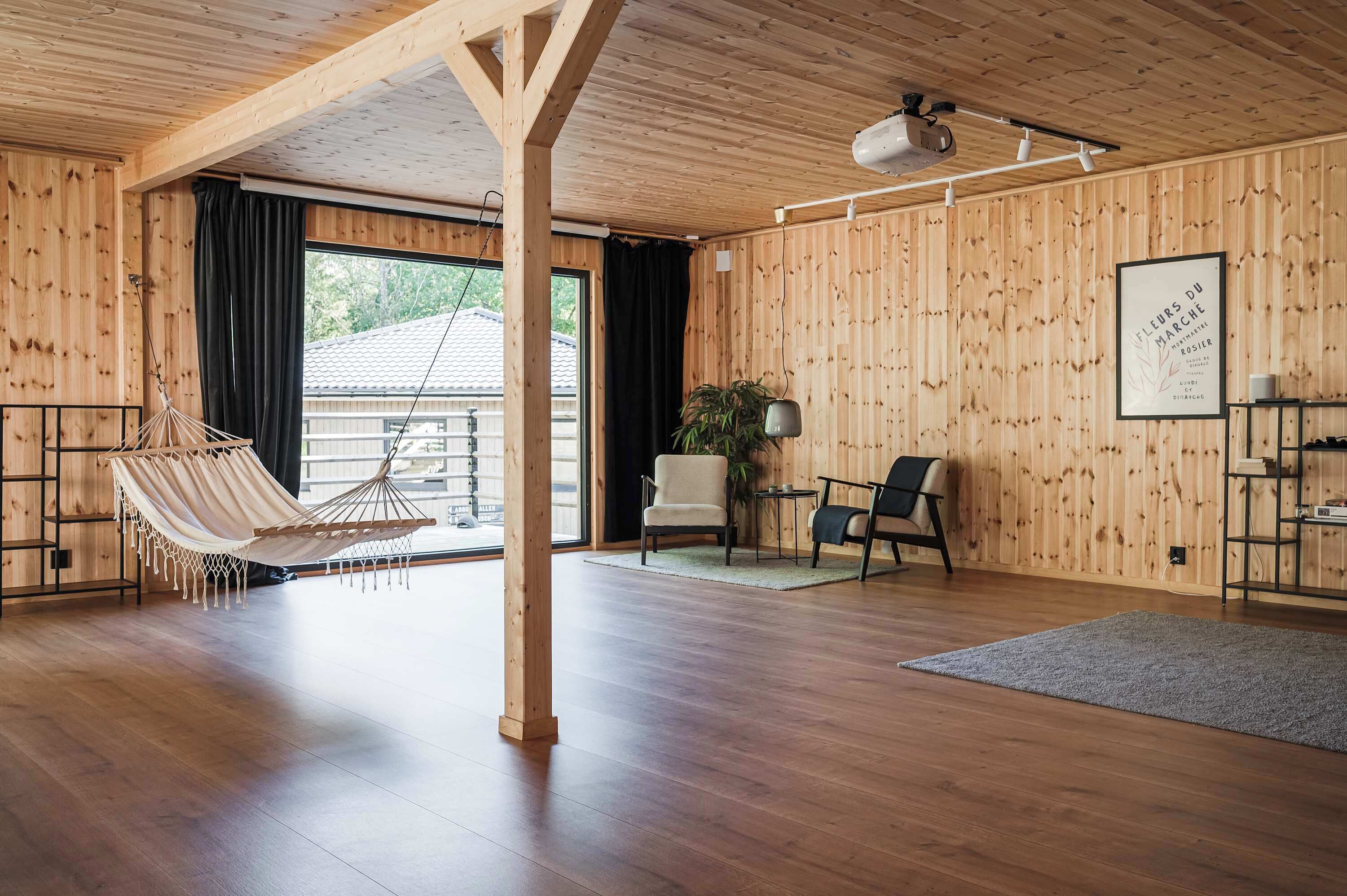 Une pièce spacieuse aux murs et au sol en bois, avec un hamac, une chaise, une petite table et un tableau encadré accroché au mur.