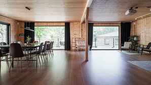 A spacious room with wooden walls and floors, a large dining table with chairs, a hanging light fixture, and a view of greenery outside. - Ladhus One - Fancy Nature Hill home by the lake (Floda)