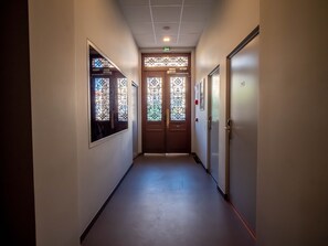 Un pasillo con una puerta de madera al final, una ventana con rejas de hierro decorativas y un detector de incendios instalado en la pared.