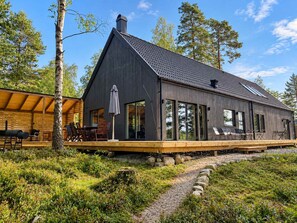 A modern cabin with a large deck, outdoor seating, and a stone foundation. - The Nabbe – Lake Escape w/ Sauna, Jacuzzi & Boats (Gällstad)