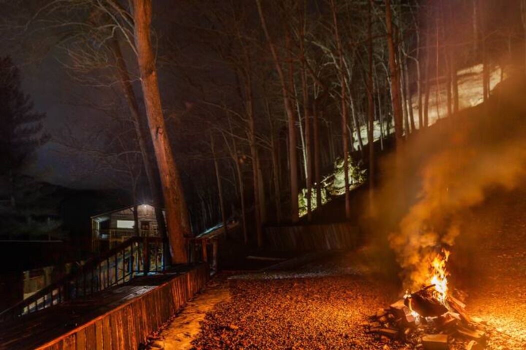 Nachtszene mit einer Holzdielen-Terrasse, einem Lagerfeuer und einem Gebäude im Hintergrund.