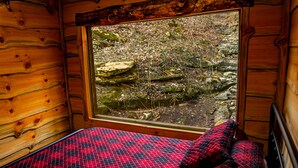 A wooden cabin room with a bed, a window with a view of rocks and vegetation. - Glamping Cabin | Huge Window | Over a Creek (Green Township)