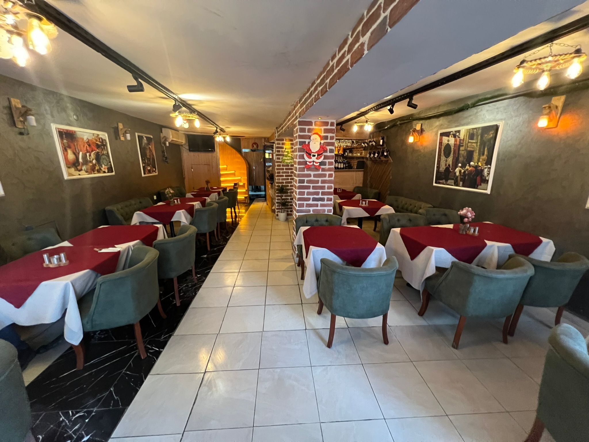 A restaurant with tables covered in red and white tablecloths, green chairs, and a brick pillar with a Santa Claus decoration.