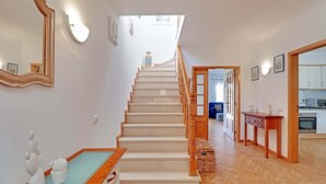 Un escalier menant à un deuxième étage, avec une rampe en bois, une petite table agrémentée d’objets décoratifs, et un coin cuisine visible au fond.