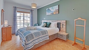Une chambre avec un grand lit, une table de chevet en bois, une commode, un luminaire suspendu, une fenêtre avec des rideaux et un tableau au mur.