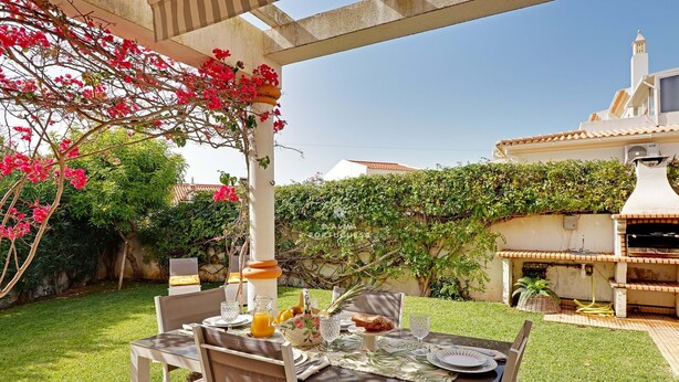 Un espace de restauration en plein air avec une table dressée pour deux personnes, entouré de verdure et agrémenté d’une pergola ornée de plantes à fleurs.