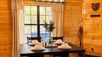 A wooden interior with a dining table set for four, a bench, and a large window with curtains.