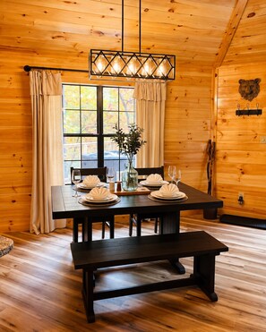 A wooden interior with a dining table set for four, a bench, and a large window with curtains.