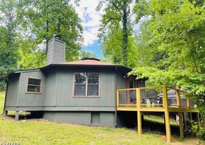 A small, single-story cabin with a chimney, a deck, and a fenced area.