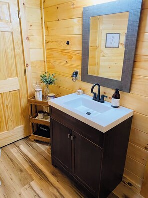 Un baño de madera con un lavamanos blanco, armarios oscuros, un espejo y una pequeña repisa con artículos de aseo.