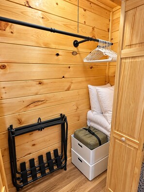 A compact room with wooden walls, a bed with white bedding, a hanging chair, and a black rack with straps.