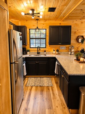 A kitchen with wooden cabinets, a stainless steel refrigerator, a granite countertop, and a window with a view of greenery.