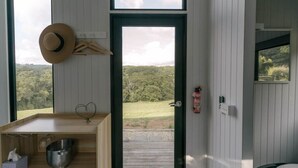 A room with a wooden desk, a hat on the wall, a fire extinguisher, and a view of a green landscape through a door. - Rainforest Sunsets by Tiny Away (Kuranda)