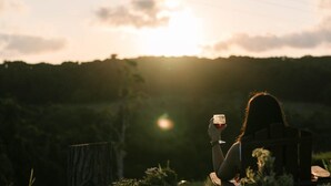 A person sitting outdoors with a drink, surrounded by greenery and hills. - Rainforest Sunsets by Tiny Away (Kuranda)