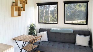 Una habitación con un sofá gris, una mesa de madera y dos ventanas con marcos negros.