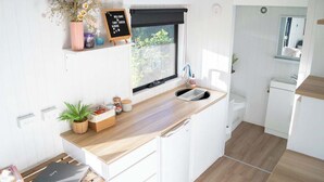 Una cocina compacta con encimera de madera, armarios blancos, una ventana con persiana negra y un pequeño letrero en una pizarra.