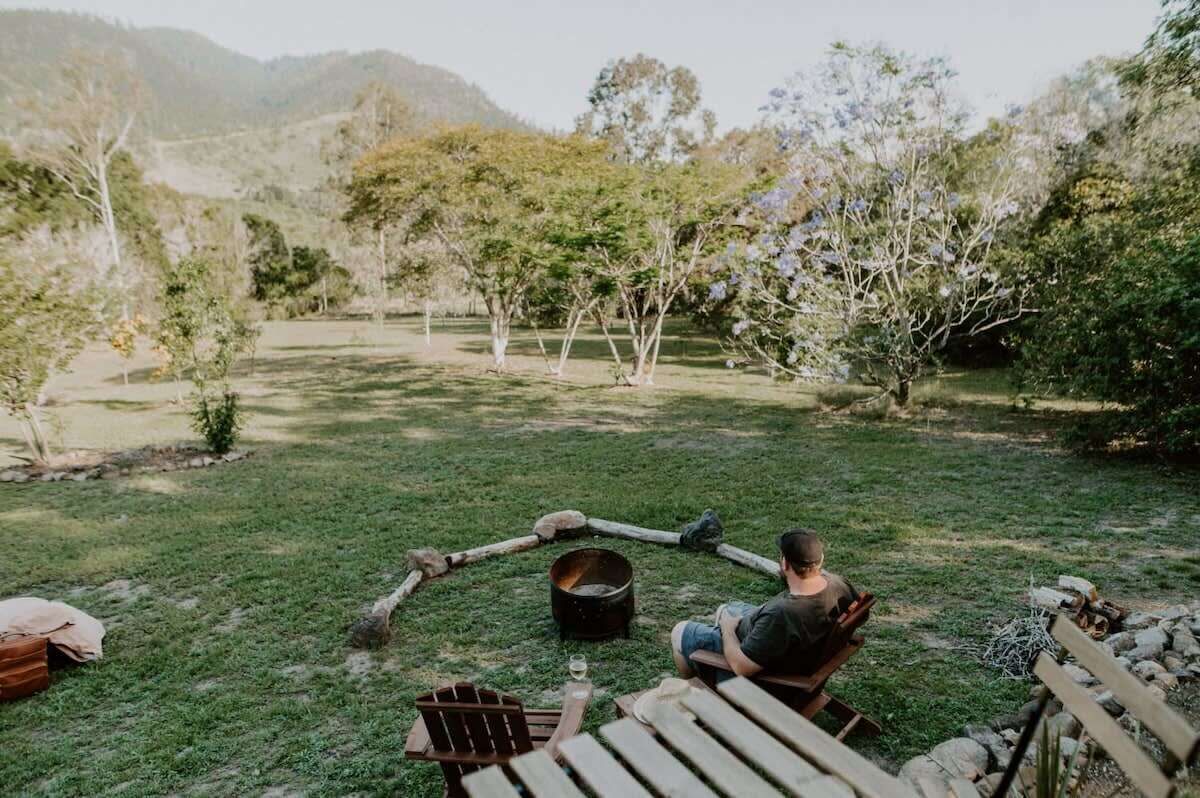 A person sitting by a fire pit in a grassy outdoor area with trees and hills in the background.