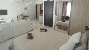 A hotel room with a large bed, a desk, a chair, a small table with a vase of flowers, and a door leading to another room. - Studio - Sleeps 2 - Balcony - 5 Min Walk To Beach (St. Paul's Bay)