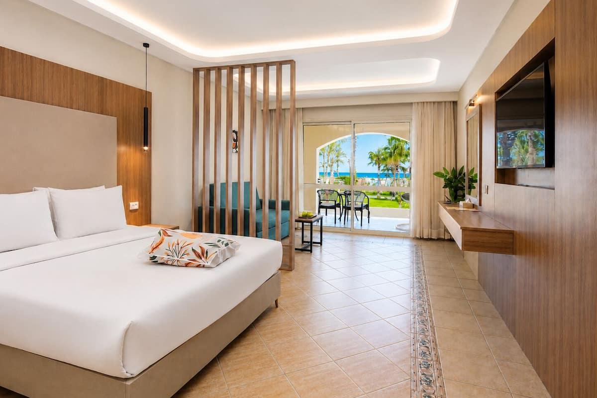 A hotel room with a bed, a TV, a view of the beach, and a balcony with outdoor furniture.