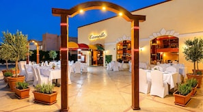 Un espace extérieur aménagé pour les repas, avec des tables et des chaises, une enseigne indiquant « Camello », et des plantes en pot.