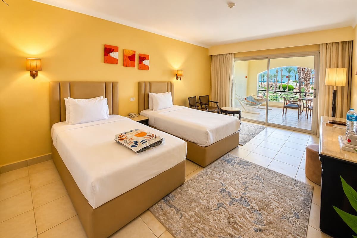 A hotel room with two beds, a desk, a chair, a small table, a lamp, and a view of the outside.