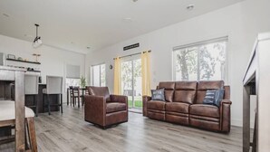 Un salón amplio con un sofá y sillón de cuero marrón, un comedor con mesa y sillas, y ventanas grandes que ofrecen vistas al exterior.