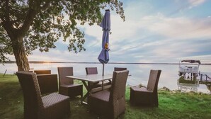 Un área de comedor al aire libre con mobiliario de mimbre y una mesa, con vista a un lago y un muelle al fondo.