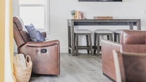 Una sala moderna con un sofá de cuero marrón, un comedor con una mesa pequeña y taburetes, un ventilador de techo y un televisor de pantalla plana montado en la pared.
