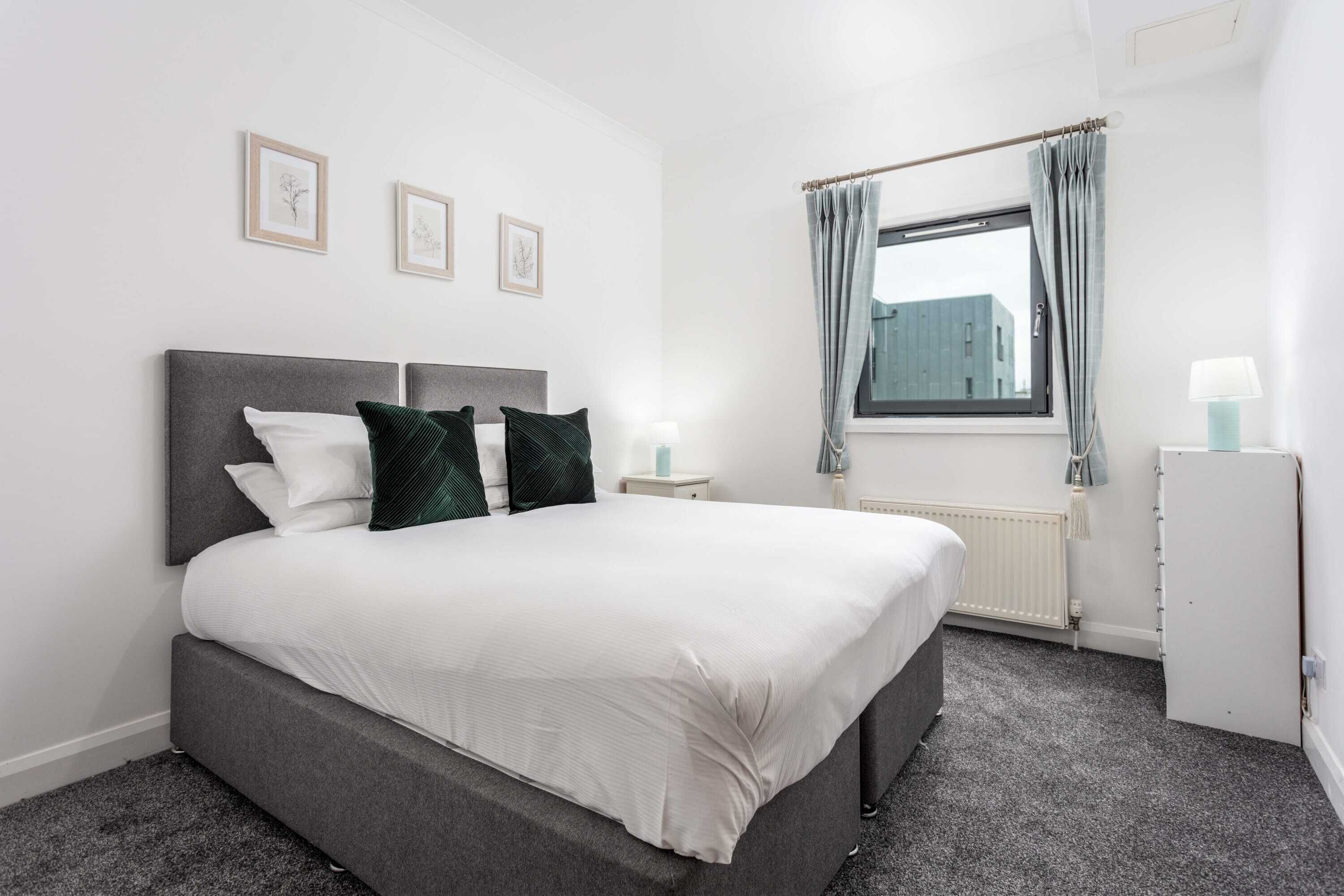 A neatly arranged bedroom with a bed, bedside tables, a window with curtains, and framed pictures on the wall.
