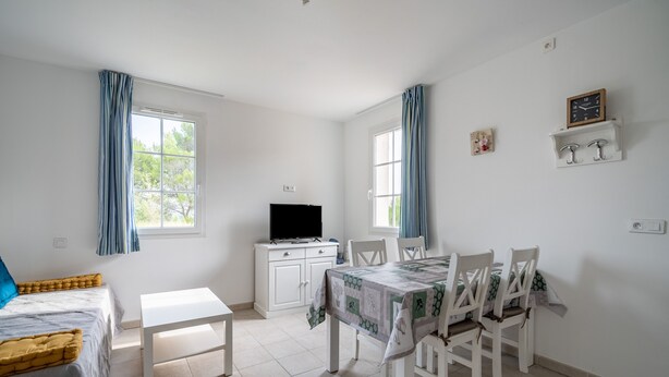 Quarto com cama, escrivaninha, mesa de jantar com cadeiras, televisão e janela com cortinas.