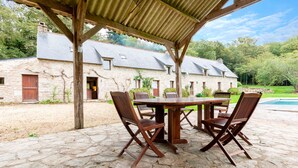 A stone patio with a wooden table and chairs, a covered area with a roof, and a stone building in the background. - Kerguestenen - Longère pour 12 avec piscine (Inguiniel)
