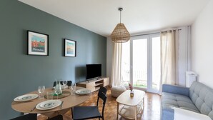 Ein Essbereich mit einem Holztisch, Stühlen und einem Fernseher auf einem Schrank. An der Wand hängen Bilderrahmen, und es gibt ein Fenster mit Vorhängen.