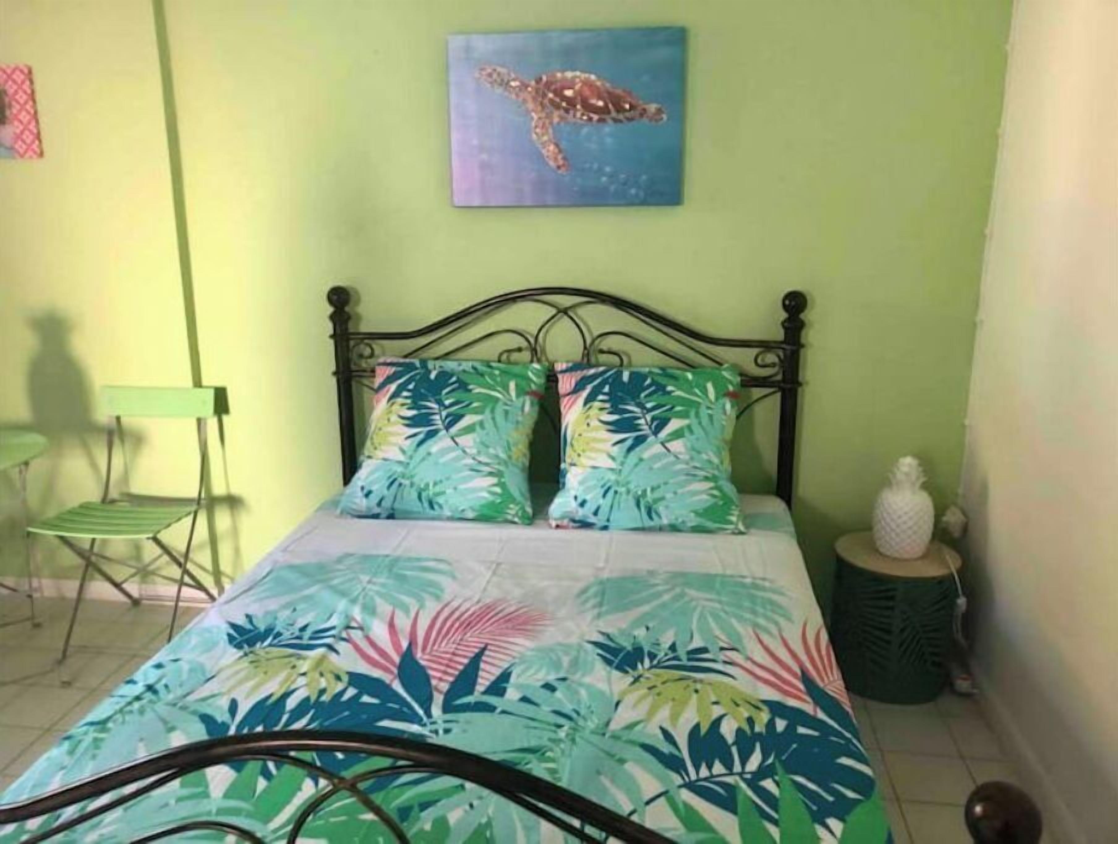 A bedroom with a bed, two pillows, a green chair, a small table with a pineapple, and a painting of a turtle on the wall.