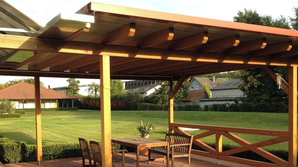 Un patio techado con una mesa y sillas de madera, vistas a un césped cuidado y una casa al fondo.