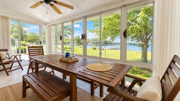 Un patio amplio con una mesa y sillas de madera, ventanas grandes que ofrecen vistas a un lago y follaje.