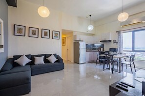 A modern living room with a dark sofa, a dining area with chairs, a kitchen with white cabinets, and a large window offering a city view.