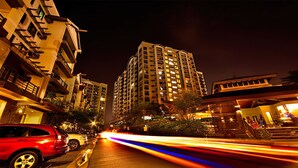 A nighttime cityscape with tall residential buildings, a red car parked on the side of the road, and light trails from moving vehicles.