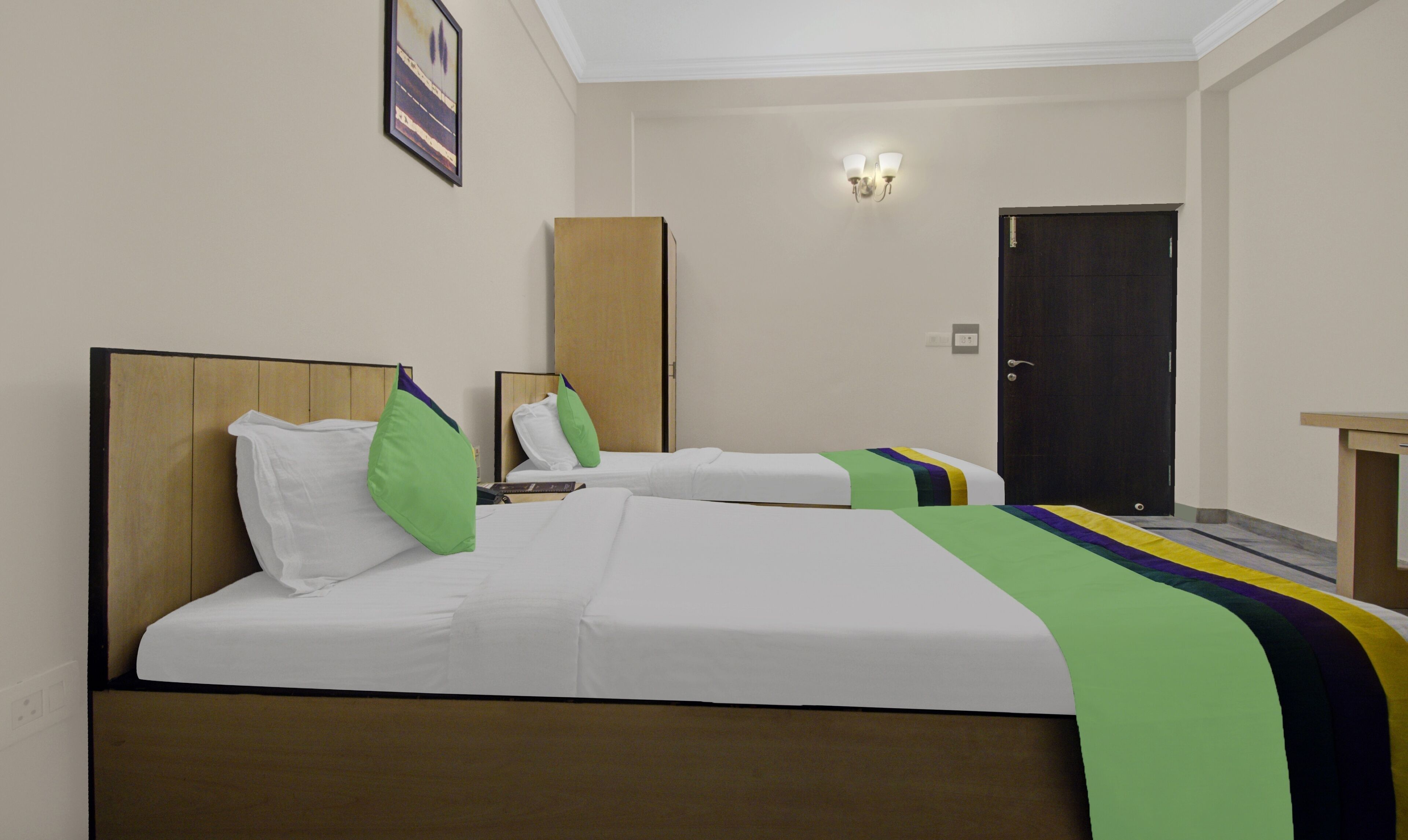 A hotel room with two beds, a wooden headboard, a bedside table, a nightstand, a door, and a wall-mounted light fixture.